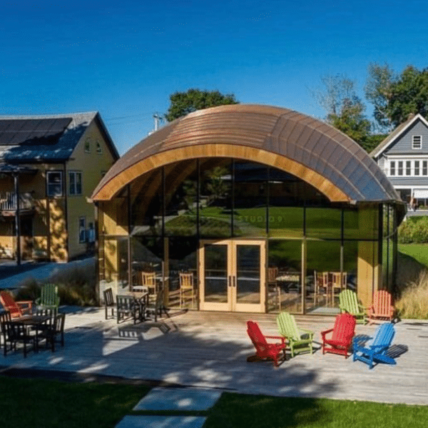 Modern arched building with glass walls and a copper roof, surrounded by colorful chairs and green lawn, adjacent to traditional houses near North Adams, Massachusetts.
