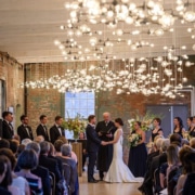 A wedding ceremony in a rustic hall with brick walls, attended by guests from North Adams hotels, watching a couple exchange vows under a canopy of string lights.