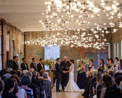 A wedding ceremony in a rustic hall with brick walls, attended by guests from North Adams hotels, watching a couple exchange vows under a canopy of string lights.