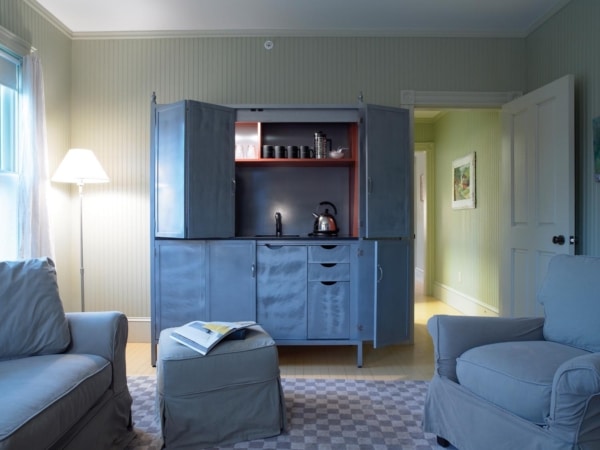 A cozy living room with soft blue sofas, an open gray cabinet displaying tea sets, a floor lamp, and a casual setup with a book on the sofa in one of the hotels near North Adams,
