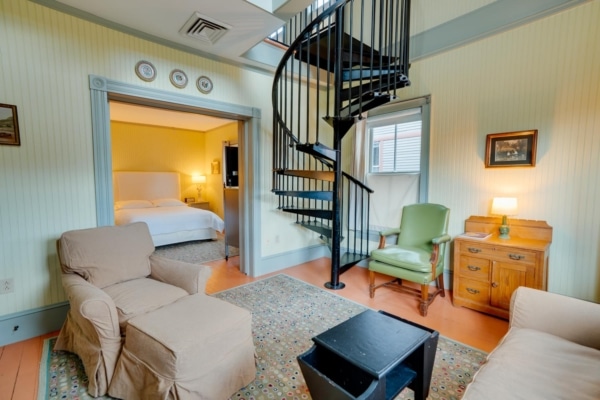 A cozy room in one of the North Adams hotels, featuring a spiral staircase, with a sitting area leading to an adjacent bedroom. The decor includes a floral rug, green chair, and soft lighting.