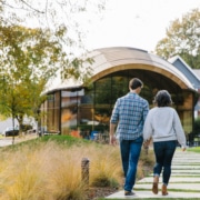 A couple walks hand-in-hand along a path leading to a modern curved building in a park-like setting near North Adams hotels with homes in the background.