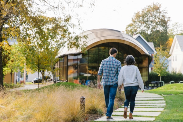 A couple walks hand-in-hand along a path leading to a modern curved building in a park-like setting near North Adams hotels with homes in the background.