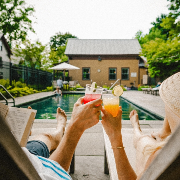 Two people relax on lounge chairs by a pool, holding up drinks for a toast; one reads a book.