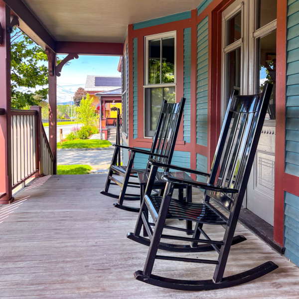Two black rocking chairs sit on a wooden porch of a blue and red house, with sunlight and greenery visible in the background.